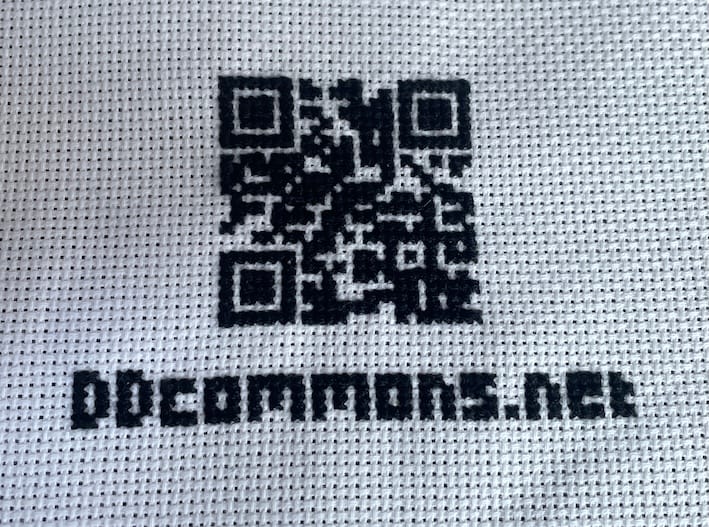 black and white cross stitch of a QR code and the text ddcommons.net